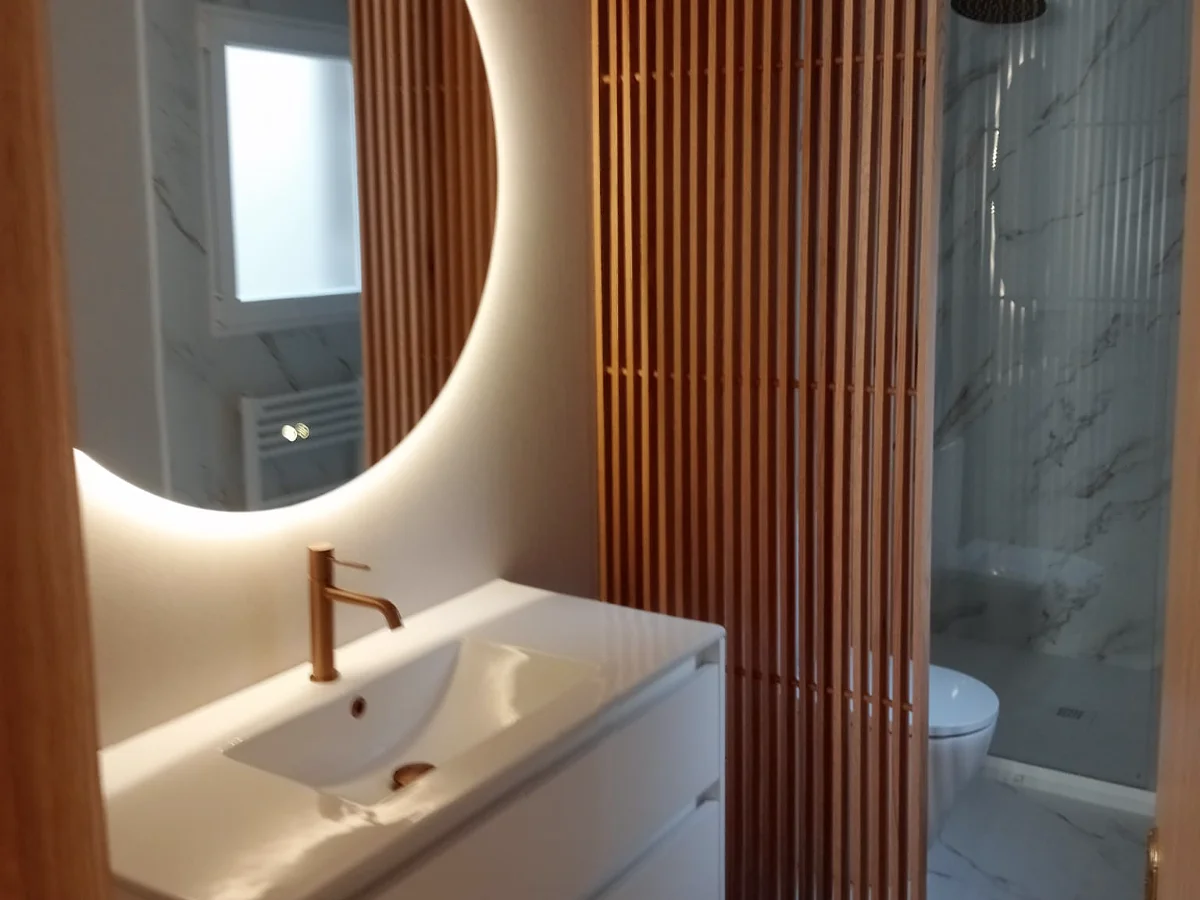 Baño con espejo redondo retroiluminado, listones de madera y ducha revestida en porcelánico tipo mármol.