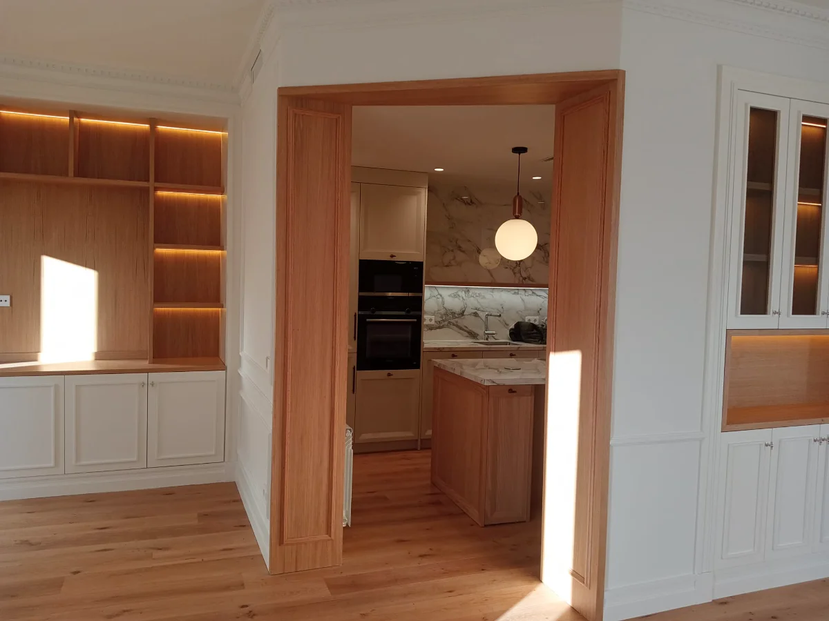 Mobiliario a medida en madera y blanco, con hornacinas iluminadas y paso enmarcado hacia la cocina.